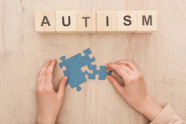 Vista parcial de las manos femeninas con rompecabezas azules en la mesa de madera cerca de cubos de madera con letras de autismo - foto de stock