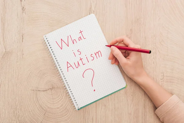 Vista recortada de la mujer escribiendo en el cuaderno lo que es cuestión de autismo en la mesa de madera - foto de stock