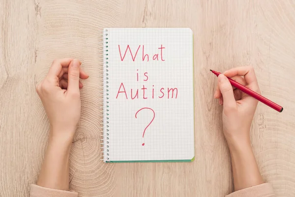 Vista recortada de la mujer sosteniendo marcador cerca del cuaderno con lo que es cuestión de autismo en la mesa de madera - foto de stock