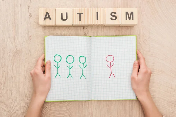 Vista recortada de la mujer sosteniendo cuaderno abierto con diferentes seres humanos dibujados cerca de cubos de madera con letras de autismo - foto de stock