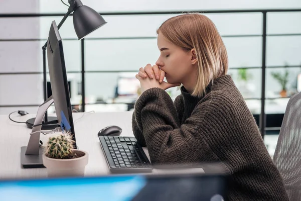 Избирательный фокус расстроенной девушки, сидящей на рабочем месте с сжатыми руками и закрытыми глазами в современном офисе — стоковое фото