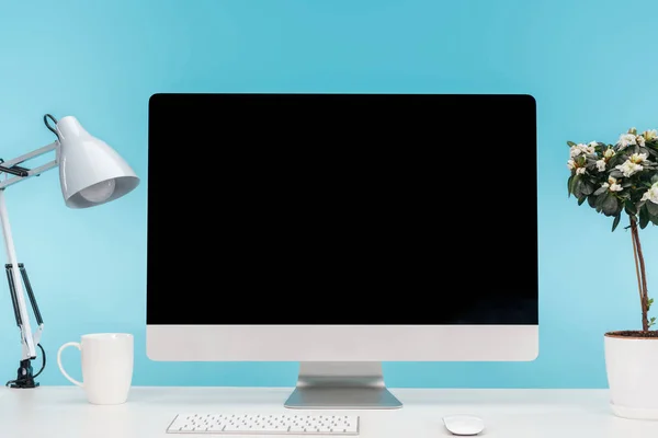 Lieu de travail avec ordinateur, lampe, tasse et pot de fleurs sur table blanche sur fond bleu — Photo de stock