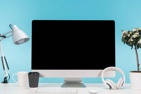 Workplace with computer, lamp, cup, coffee to go, headphones and flowerpot on white table on blue background — Stock Photo