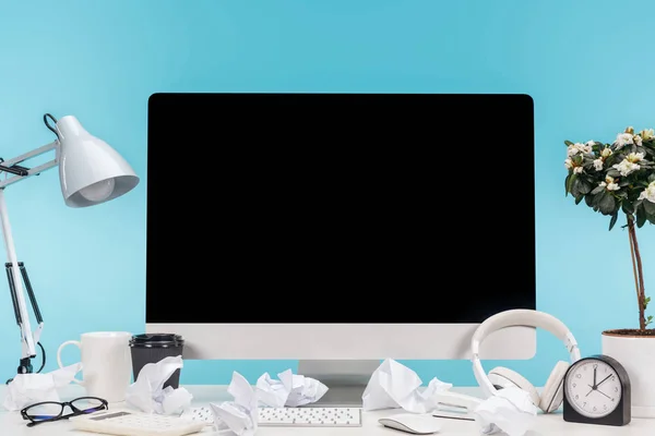 Lugar de trabajo desordenado con computadora, lámpara, taza, café para llevar, papeles arrugados, auriculares y maceta en la mesa blanca sobre fondo azul - foto de stock