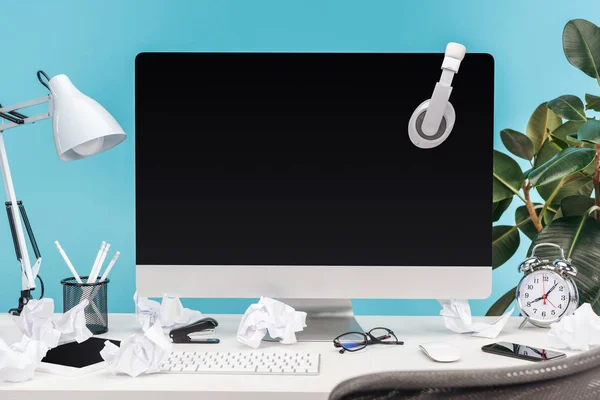 Messy workplace with headphones on computer, lamp, crumpled papers and stationery on white table on blue background — Stock Photo