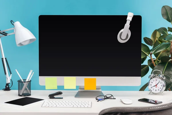 Рабочее место с наушниками и пустыми липкими нотами на мониторе компьютера, лампой и канцелярскими принадлежностями на белом столе на синем фоне — стоковое фото