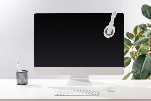 Workplace with headphones on computer monitor and stationery on white table near green plant on grey background — Stock Photo