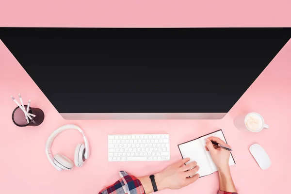 Cropped view of man writing in notebook near computer on pink background — Stock Photo