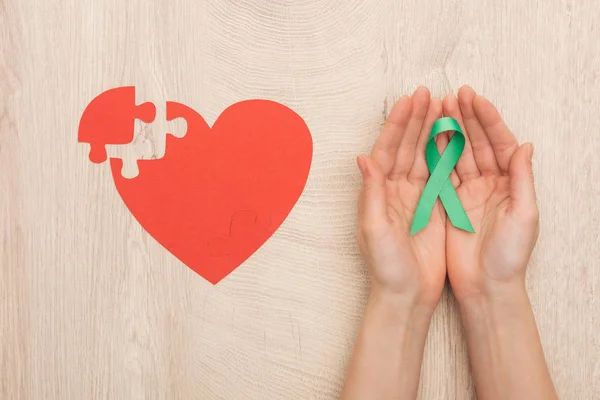 Partial view of woman holding green ribbon and paper heart on wooden background — Stock Photo