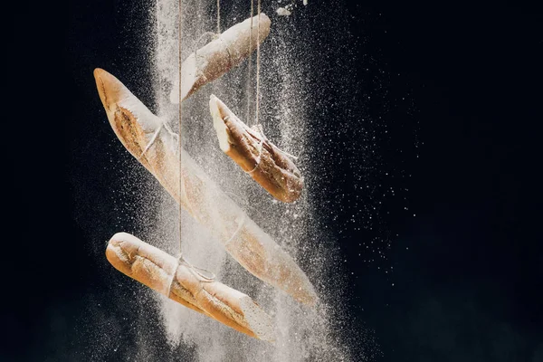 White flour falling at loaves of fresh baked baguettes on ropes on black background — Stock Photo