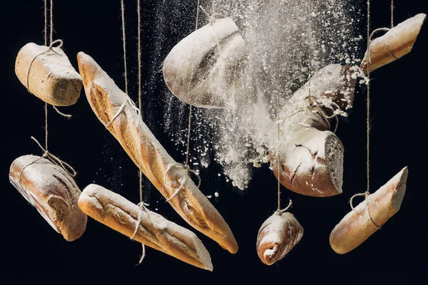 Farinha branca caindo em pão fresco assado pendurado em cordas em fundo preto — Fotografia de Stock