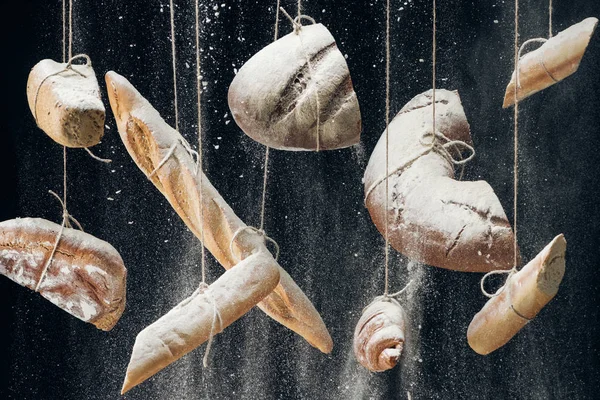 White flour falling at baked bread, baguettes and croissant hanging on ropes on black background — Stock Photo