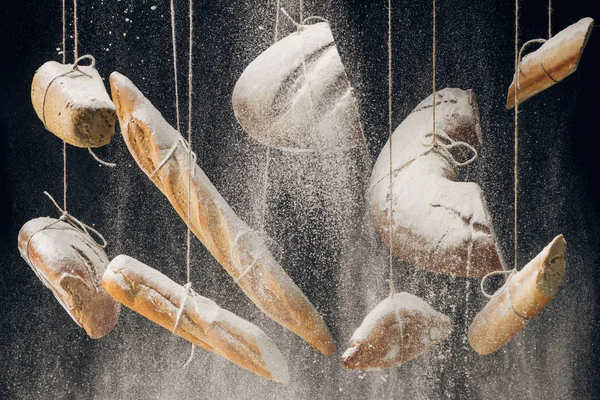 Farinha branca caindo em pão fresco assado, baguetes e croissant pendurado em cordas em fundo preto — Fotografia de Stock