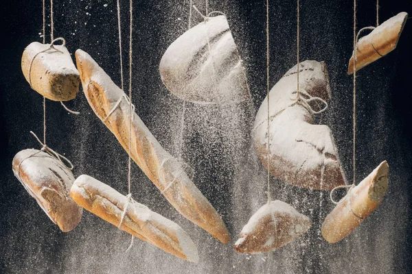 Farine blanche tombant au pain frais cuit au four, baguettes et croissant accroché aux cordes sur fond noir — Photo de stock