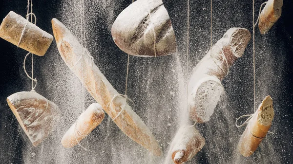 Mehl fällt auf gebackenes Brot, Baguettes und Croissants, die an Seilen auf schwarzem Hintergrund hängen — Stockfoto