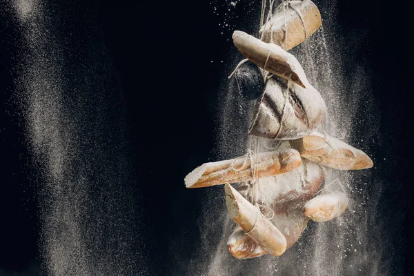 Harina blanca cayendo en pan, baguettes y croissant sobre cuerdas sobre fondo negro - foto de stock