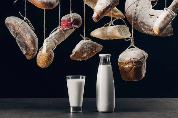 Latte in vetro e bottiglia su tavola e pane con farina appesa su corde isolate su nero — Foto stock
