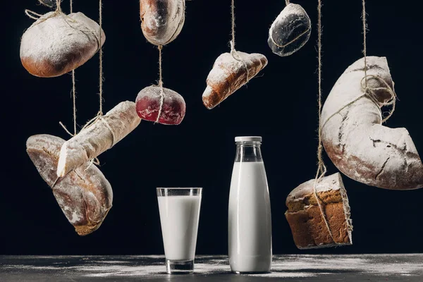 Leche en la mesa y pan con harina colgando de cuerdas aisladas en negro - foto de stock