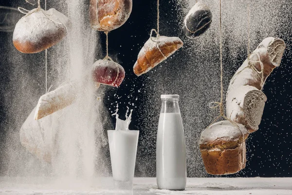 Milk splashing in glass on table and four falling on bread hanging on strings isolated on black — Stock Photo