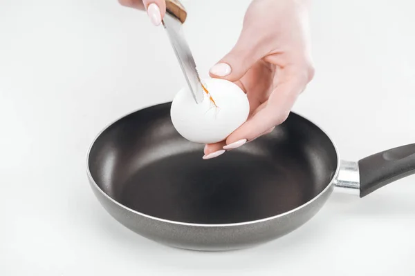 Частковий вигляд жінки, що розбиває яйце на сковороді з ножем на білому тлі — стокове фото
