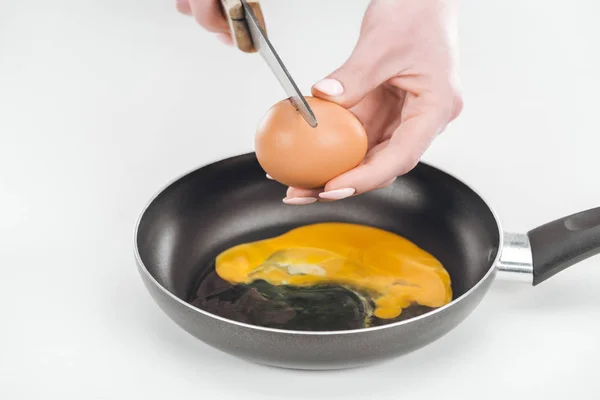 Vue recadrée d'une femme écrasant un œuf de poulet avec un couteau tout en préparant des œufs brouillés dans une casserole sur fond blanc — Photo de stock