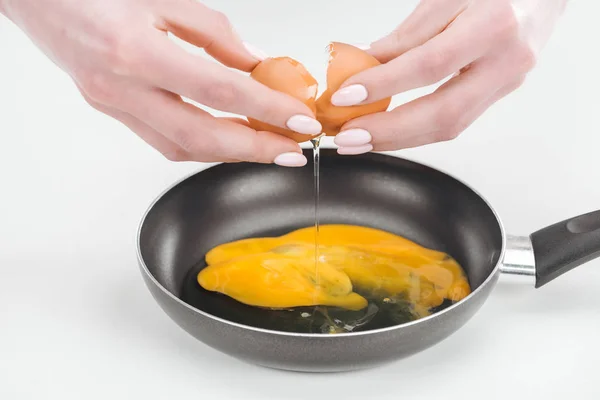 Vista recortada de la mujer rompiendo huevo de pollo fresco mientras prepara huevos revueltos en la sartén sobre fondo blanco - foto de stock