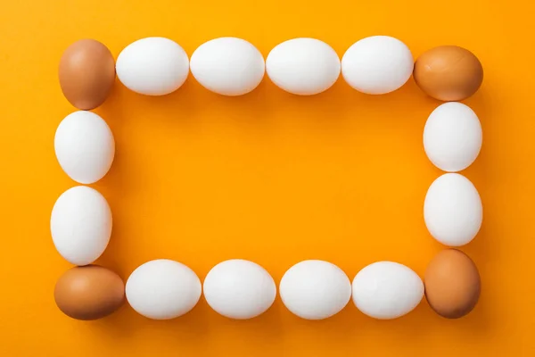 Vue de dessus des oeufs de poulet biologiques blancs et bruns entiers disposés en cadre carré sur fond orange vif avec espace de copie — Photo de stock