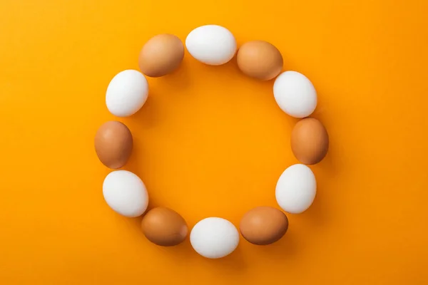 Vue de dessus des oeufs de poulet biologiques blancs et bruns entiers disposés en cadre rond sur fond orange vif avec espace de copie — Photo de stock