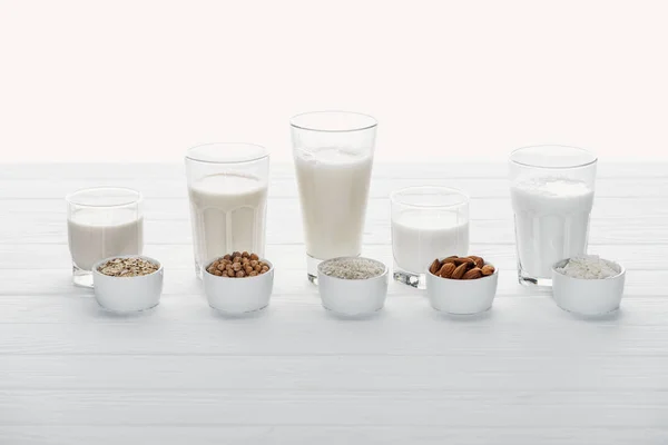 Стаканы с кокосом, горохом, овсянкой, рисом и миндальным молоком с ингредиентами в мисках, изолированных на белом — стоковое фото