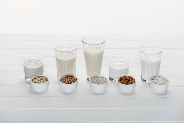 Vasos con coco, garbanzo, avena, arroz y leche de almendras sobre mesa de madera blanca con ingredientes en cuencos aislados sobre blanco - foto de stock