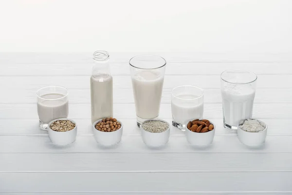 Vasos y botella con coco, garbanzo, avena, arroz y leche de almendras en la superficie de madera blanca con ingredientes en cuencos aislados en blanco - foto de stock