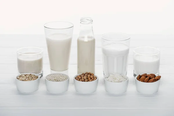 Vasos y botella con coco, garbanzo, avena, arroz y leche de almendras en mesa de madera blanca con ingredientes en cuencos aislados en blanco - foto de stock