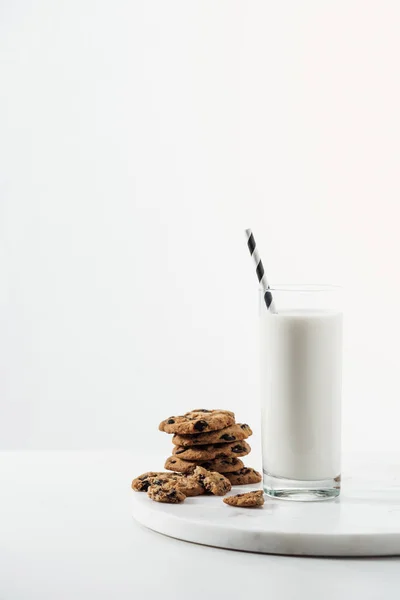 Vaso con leche fresca y paja cerca de galletas de chocolate en soporte de mármol aislado en blanco - foto de stock