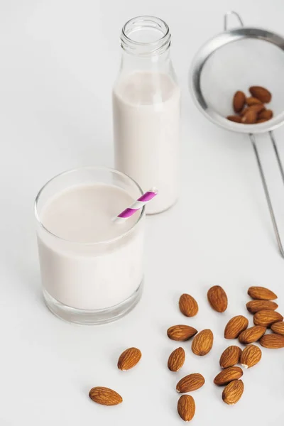 Latte di mandorla fresco in bottiglia e bicchiere con cannuccia vicino a mandorle sparse e setaccio — Foto stock