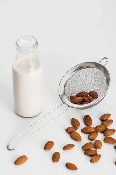 Vegan almond milk in bottle near scattered almonds and sieve on grey background — Stock Photo