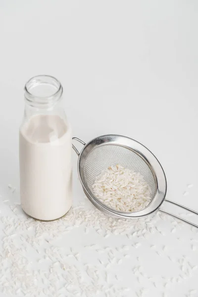 Latte di riso in bottiglia vicino a riso sparso e setaccio su fondo grigio — Foto stock