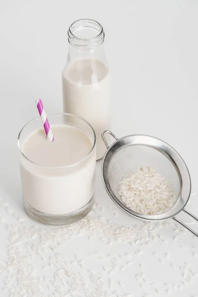 Rice milk in bottle and glass with straw near scattered rice and sieve on grey background — Stock Photo