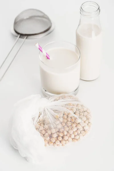 Chickpea in white cheesecloth near bottle and glass with vegan chickpea milk and sieve on grey background — Stock Photo