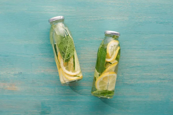Вид сверху на детоксикационный напиток в бутылках с ломтиками лимона и огурца, листья мяты на голубой деревянной поверхности — стоковое фото