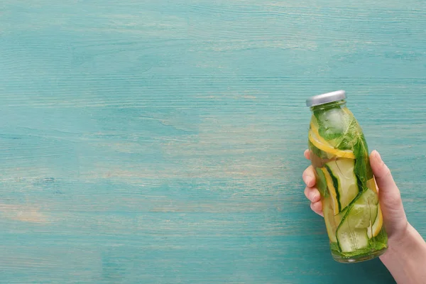 Cropped view of woman holding bottle with detox drink on blue wooden surface — Stock Photo