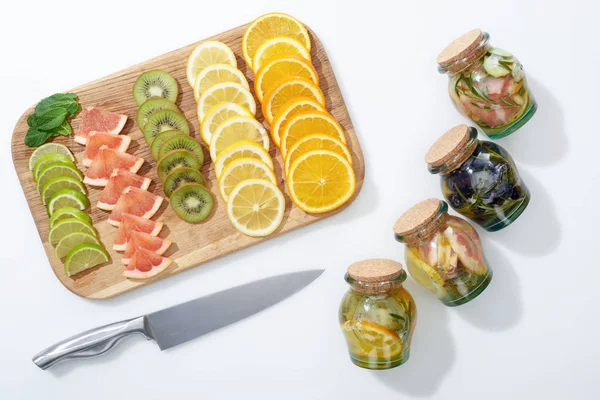 Vista superior de bebidas desintoxicantes en frascos cerca de cuchillo y rebanadas de fruta en tabla de cortar de madera - foto de stock