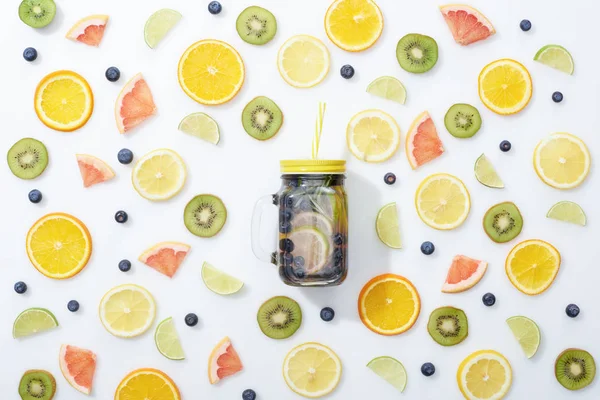 Vue du dessus de la boisson de désintoxication en pot avec de la paille parmi les fruits tranchés et les bleuets sur fond blanc — Photo de stock