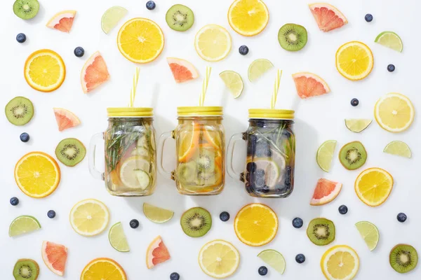 Top view of detox drinks in jars with straws among sliced fruits and blueberries on white background — Stock Photo