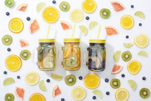 Vue de dessus des boissons de désintoxication dans des bocaux avec des pailles parmi les fruits tranchés et les bleuets sur fond blanc — Photo de stock