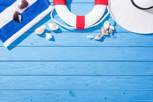 Vista superior de toalha listrada, óculos de sol, bóia salva-vidas, chapéu flexível e conchas no fundo de madeira azul com espaço de cópia — Fotografia de Stock