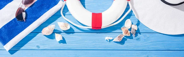 Colpo panoramico di asciugamano a righe, occhiali da sole, salvagente, cappello floppy bianco e conchiglie su sfondo blu in legno — Foto stock