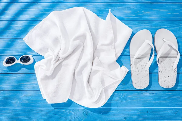 Draufsicht auf weißes zerknülltes Handtuch, Sonnenbrille und Flip-Flops auf blauem Holzhintergrund — Stockfoto