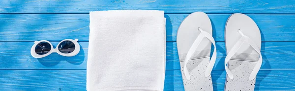 Panoramic shot of white folded towel, retro sunglasses and flip flops on blue wooden background — Stock Photo