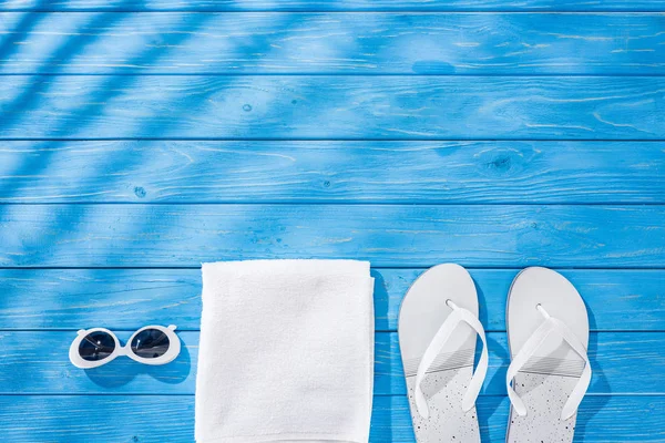 Vista superior de toalha branca dobrada, óculos de sol retro e chinelos em fundo de madeira azul com sombras e espaço de cópia — Fotografia de Stock