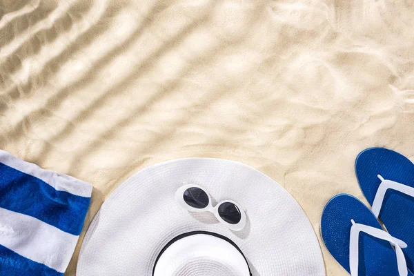 Vista superior de toalla rayada, gafas de sol retro, sombrero floppy y chanclas azules en la arena con sombras y espacio de copia - foto de stock